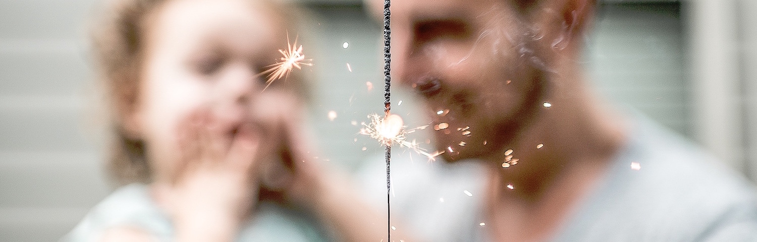 closeup of sparkler with dad holding toddler out of focus in the background