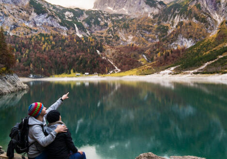a couple sitting on a log at the edge of a lake pointing up to the scenery mountains
